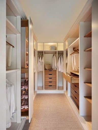 A long walk-in closet with classic wood shelving on either side, leading to a lit space with drawers and hanging areas.




