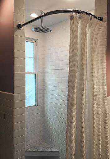 A small bathroom shower with a curved shower rod and beige curtain partially drawn, showcasing a window. 