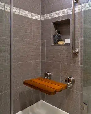 Tiny shower space with Folding or Telescoping Bench textured grey tiles, a recessed shelf, and a wooden corner seat, exemplifying efficient use of space.