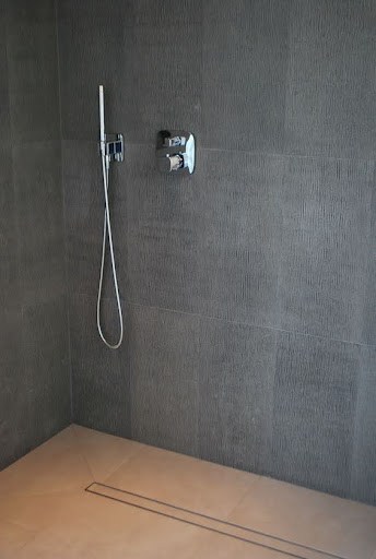 Small shower area with a Linear Drain and simple hand-held shower fixture against a dark tiled wall, showcasing understated design.